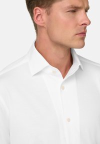 Uomo che indossa una camicia bianca con colletto a punta, mostrato dalla gola fino a metà torace, che guarda a destra.