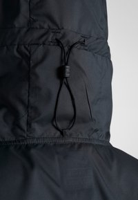 Primer plano de la capucha de una chaqueta negra con cordón elástico ajustable y tope de plástico para apretar.