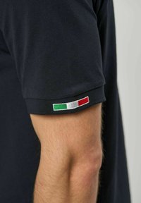 Kortermet svart skjorte på arm, med et lite brodert italiensk flagg på ermekanten.