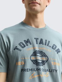 Gaiši zils kokvilnas T-kreklis ar apaļu kaklu, ar tumši zilu un oranžu drukātu tekstu, logo un apļveida dizainu uz priekšpuses.