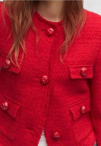 Veste rouge texturée avec quatre poches à rabat, grands boutons brillants et col arrondi. Le tissu semble tissé et chaud.