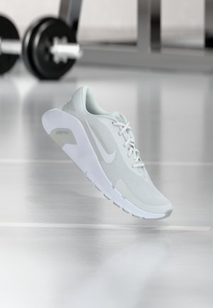 Chaussure de running Nike gris clair suspendue au-dessus d'un sol de salle de sport avec des poids et du matériel flous en arrière-plan.