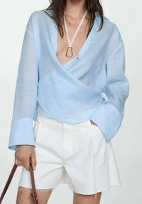 Femme portant un haut enveloppant léger bleu clair et un short taille haute blanc, tenant un sac à main marron d'une main, avec l'autre main dans la poche.