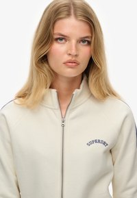 Sweatshirt crème à fermeture éclair avec col et poignets côtelés. Arborant un logo brodé bleu marine. Texture lisse et conception à manches raglan.