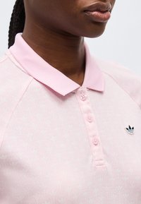 Κοντινό πλάνο ατόμου που φοράει ανοιχτό ροζ πόλο με γιακά, κουμπιά και μικρό σκούρο μπλε λογότυπο Adidas στο στήθος.