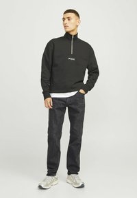 Sudadera de forro polar negra con cuello de media cremallera y logo blanco, combinada con pantalones vaqueros oscuros y zapatillas claras, de pie frente a un fondo liso.
