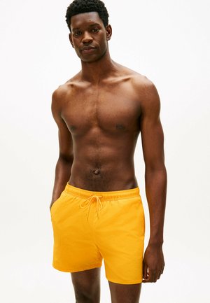 Młody, muskularny mężczyzna stojący z rękami w kieszeniach, ubrany w jasne żółte kąpielówki, na jednolitym białym tle.