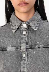 Veste en denim gris avec col, ornée de clous métalliques et de boutons-pression. La vue rapprochée met en avant la texture et les détails du tissu.