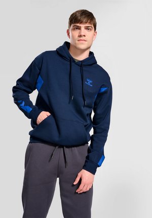 Jeune homme portant un sweat à capuche bleu marine avec des détails en chevrons bleus et un pantalon de survêtement gris foncé, debout avec une main dans la poche.