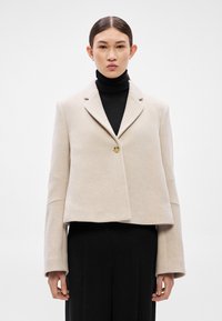 Μπεζ cropped σακάκι με γιακά με χαραγή, κλείσιμο με ένα κουμπί και φαρδιά μανίκια, φορεμένο πάνω από μαύρο ζιβάγκο και μαύρο παντελόνι.