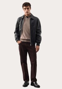 Czarna skórzana kurtka bombowa na beżowym swetrze zapinanym na zamek, w połączeniu z ciemnobrązowymi spodniami i czarnymi butami, stojąca na neutralnym tle.