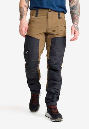 Man wearing two-tone brown and black cargo-style pants and black hiking shoes with red laces, standing on white background.