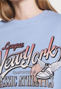 Ljusblå tröja med rund hals som har en stark grafisk tryck med "League New York" och en design av en bevingad sneaker. Mjuk bomullsmaterial.