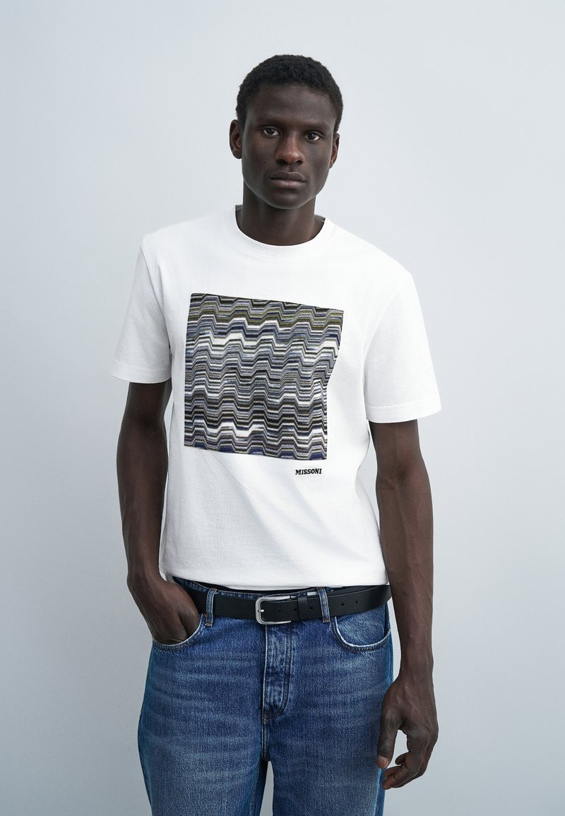 Νέος άνδρας που φοράει λευκό T-shirt με πολύχρωμο ζιγκ-ζαγκ μοτίβο και μπλε τζιν, στέκεται μπροστά σε απλό ανοιχτό γκρι φόντο.