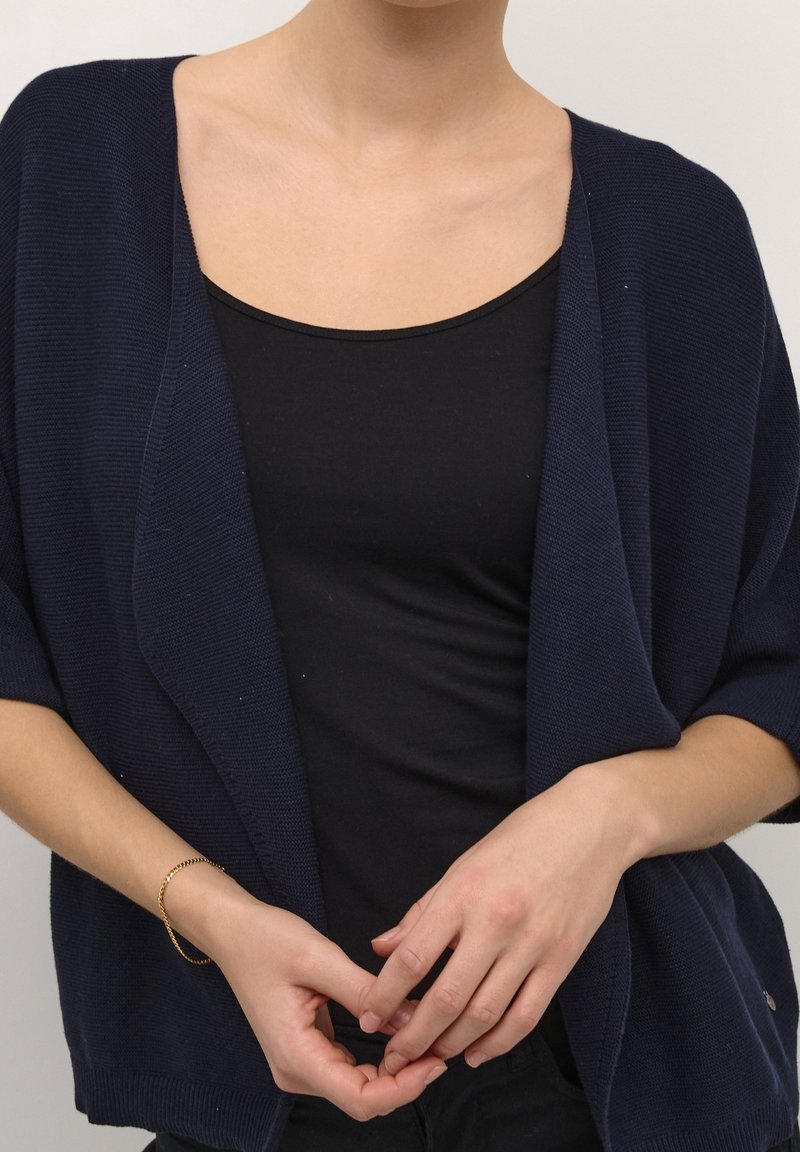 Gilet en maille lâche de couleur marine avec des manches larges, porté sur un haut noir, présentant une coupe décontractée et un tissu texturé. Bracelet en or au poignet.