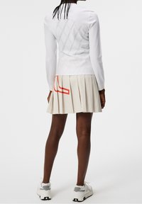 Persona vistiendo una blusa larga blanca de textura, falda plisada beige con gráfico rojo, zapatillas blancas y grises, de pie contra un fondo liso.
