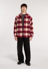 Ung mand står iført en rød- og beige-rutede skjorte med knapper, sorte bukser, sorte sko og en mørkegrå t-shirt.