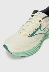 Zapatilla de running color beige claro con detalles en verde, parte superior de malla transpirable, suela texturizada y cordones planos. Diseño de suela ligeramente curvada.