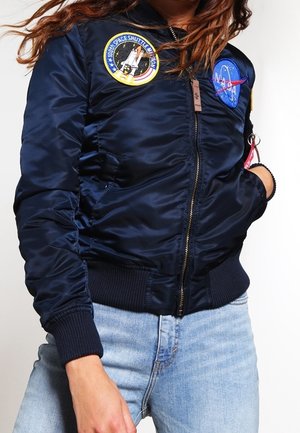 Woman wearing a navy satin bomber jacket with NASA and space shuttle mission patches, paired with light blue jeans.