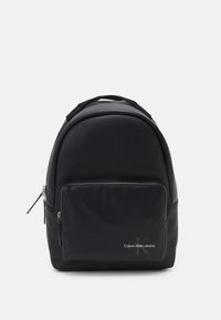 Zaino nero con una texture liscia, design arrotondato e dettagli in metallo argentato. Presenta una tasca frontale e il logo "Calvin Klein Jeans" nella parte inferiore.