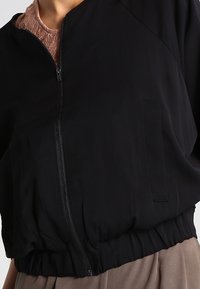 Svart zip-jacka med elastisk nederkant som bärs över en rosa spetsblus och beige byxor, som visar överkropp och nacke.