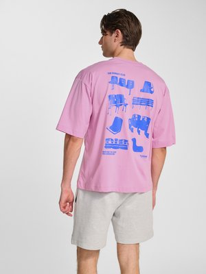 Mand iført lyserød oversized t-shirt med blå grafikprint af forskellige stol design og lysegrå shorts, vendt væk fra seeren.