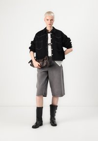 Blazer preto cropped com costura branca em contraste, calções cinzentos oversized e botas altas pretas até ao joelho. Modelo a segurar uma mala castanha.