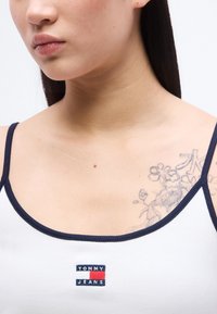 Νεαρό άτομο που φοράει λευκό αμάνικο με μπλε περίγραμμα και λογότυπο Tommy Jeans, εμφανίζοντας ένα λουλουδάτο τατουάζ στον αριστερό ώμο.