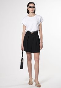 T-shirt bianca a maniche corte, pantaloni shorts neri a vita alta con pieghe, fibbia della cintura circolare dorata, occhiali da sole trasparenti, décolleté beige open-toe, borsa nera.