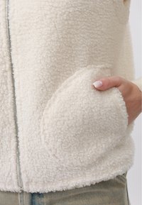 Main avec des ongles manucurés dans la poche d'une veste polaire texturée couleur crème avec une fermeture éclair argentée et un jean en denim clair.