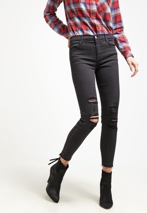 Personne portant un jean skinny noir déchiré, des bottines noires et une chemise à carreaux rouge et bleue, se tenant avec une jambe légèrement pliée.