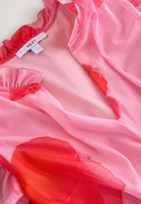 Átlátszó rózsaszín felső fodros nyakkivágással, vörös akvarell mintával. Az anyag könnyűnek és puhanak tűnik, textúrát adva. A címke "NEXT" feliratú.