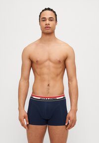 Boxers bleu foncé avec une ceinture élastique blanche et rouge, présentant un détail de logo. Le matériau semble lisse et extensible. Modèle debout avec une expression neutre.