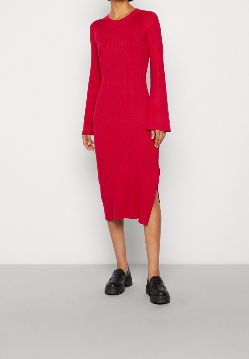 Rotes geripptes Strickkleid mit langen Glockenärmel, Midilänge, seitlichen Schlitzen, kombiniert mit schwarzen klobigen Loafers. Glatte Textur, einfarbiges Design.