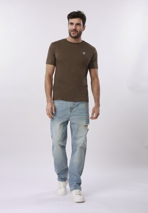 Man walking wearing a brown short-sleeve t-shirt, light blue ripped jeans, and white sneakers on a plain light background.