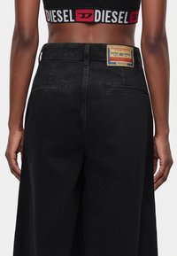 Πίσω όψη ατόμου που φοράει μαύρο crop top με σήμα Diesel και μαύρο τζιν ψηλόμεσο με ετικέτα Diesel Industry στη μέση.