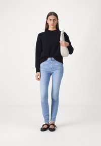 Schwarzer Strickpullover, hellblaue High-Waist-Jeans mit Knopfverschluss vorne und schwarze flache Schuhe. Das Modell trägt eine cremefarbene Umhängetasche.