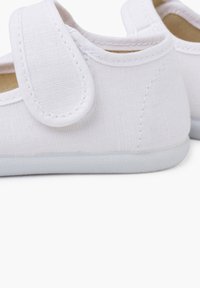 Scarpe da ginnastica bianche in tela con cinturino in velcro, dotate di suola in gomma piatta e tessuto testurizzato, progettate per comfort e facilità di indossare.