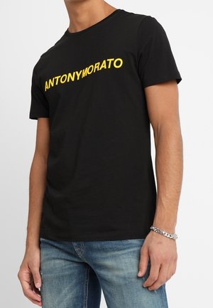 Mężczyzna noszący czarną koszulkę z żółtym napisem "ANTONY MORATO", jasnoniebieskie dżinsy i srebrną bransoletkę na prawym nadgarstku.