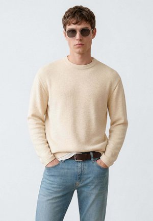 Junger Mann trägt einen beigen Strickpullover, hellblaue Jeans, braunen Gürtel und Sonnenbrille, steht mit den Händen in den Taschen vor einem einfarbigen Hintergrund.