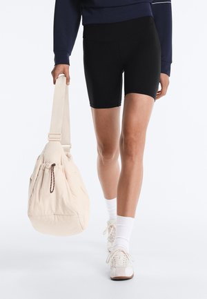 Oseba, ki nosi črne kolesarske kratke hlače, bele nogavice, športne copate, mornarsko modro majico in v roki drži bež torbo z vrvico za zapiranje na paščku.