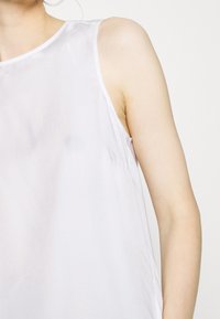 Mujer con una blusa sin mangas de color lavanda claro y escote redondo, mostrada desde los hombros hasta la mitad del brazo sobre un fondo blanco liso.