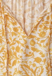 Vêtement à encolure en V avec un motif floral jaune sur tissu blanc, montrant des coutures détaillées et un tissu intérieur avec une version délavée de l'imprimé.