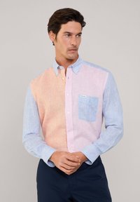 Camisa de botones con diseño a rayas en secciones de color naranja pastel y azul. El bolsillo izquierdo es de un azul claro con una pequeña etiqueta.