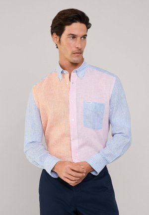 Chemise à boutons avec un design rayé comportant des sections en orange pastel et bleu. La poche gauche est bleue clair avec une petite étiquette.