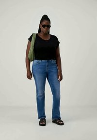 Črn majica s kratkimi rokavi, modri ravni kavbojke, zelena teksturirana torba, črne sandale in sončna očala. Enostavna svetla podlaga.