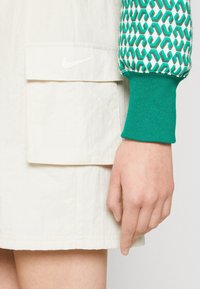 White jacket with a green patterned sleeve, featuring a ribbed cuff. Includes a front pocket with a small white logo on the chest.