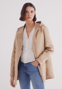 Giacca a cappuccio beige chiaro con una superficie liscia, bottoni a pressione e una vestibilità morbida. Indossata sopra una maglietta bianca e jeans in denim.
