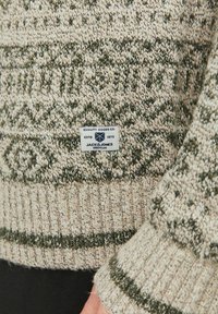 Beige und grüner gestrickter Pullover mit Mustern, gerippten Bündchen und einem Etikett mit Markeninformationen. Die Textur wirkt weich und gemütlich.