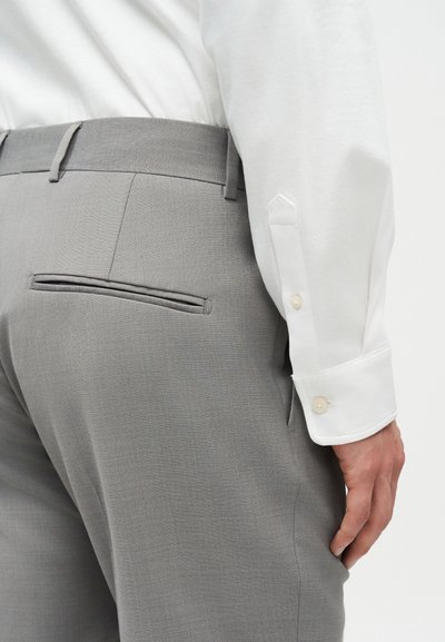 Προσαρμοσμένο ανοιχτό γκρι παντελόνι με πίσω τσέπη welt, φορεμένο με λευκό πουκάμισο μακρυμάνικο, με κουμπωμένο μανίκι και χέρι στο πλάι.